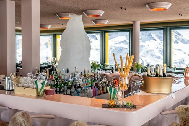 Innenraum des White Marmot mit mehreren gedeckten Tischen und Sitzplätzen mit Fellauflagen. Auf den Tischen stehen Gläser, Teller, Besteck, Servietten, Gewürzmühlen und Vasen mit Tulpen. Im Hintergrund befindet sich eine Bar mit zahlreichen Gläsern und Getränkeflaschen. In der Mitte des Raumes steht eine grosse weisse, skulpturartige Figur. Durch die grossen Fenster sind Teile der umliegenden Gebäude und eine verschneite Fläche zu sehen. An der Decke hängen mehrere runde Leuchten mit orangefarbenen Innenflächen