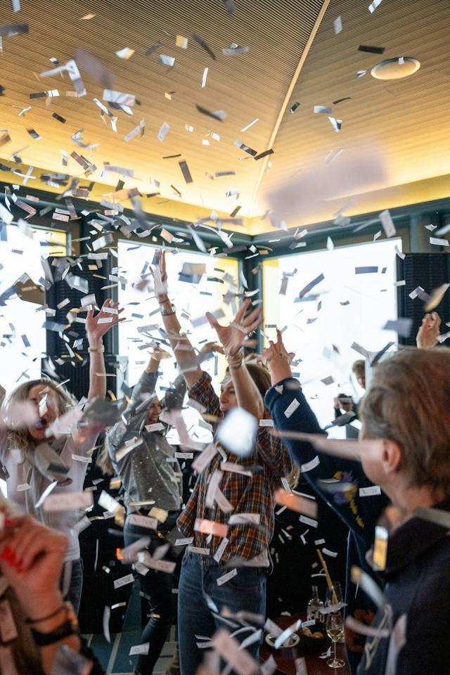 Mehrere Personen feiern beim Lunch & Dance im Restaurant White Marmot und strecken ihre Hände in die Luft, während zahlreiche schwarze, weisse und farbige Papierstreifen durch den Raum fliegen. Im Hintergrund sind grosse Fenster, dunkle Vorhänge und eine holzverkleidete Decke zu sehen. Im unteren Bildbereich stehen Tische mit Gläsern und Getränken.