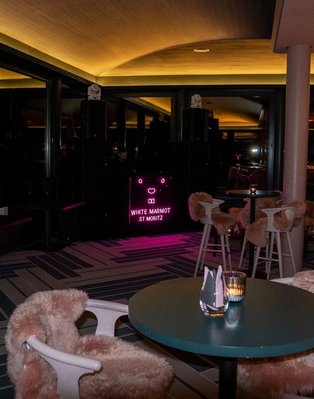Innenansicht des White Marmot St. Moritz mit DJ-Pult und Lautsprechern im Hintergrund. Das beleuchtete DJ-Pult trägt den Schriftzug «WHITE MARMOT ST MORITZ». Im Vordergrund stehen runde Tische mit Kerzen sowie weisse Stühle und Barhocker mit Fellauflagen. Der Raum ist mit grossen Spiegel- und Fensterflächen, einer holzverkleideten Decke und farbiger Beleuchtung ausgestattet.