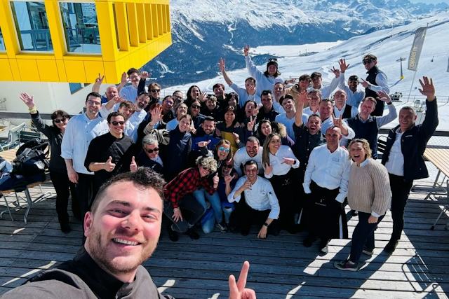 Gruppenfoto von rund 30 Mitarbeitern des Restaurants White Marmot auf der Sonnenterrasse vor dem gelben Gebäude der Bergstation Corviglia. Die Personen stehen und sitzen dicht beieinander. Viele von ihnen heben die Hände oder zeigen mit den Fingern in Richtung Kamera. Im Vordergrund hält eine Person die Kamera für ein Selfie bereit. Im Hintergrund sind eine verschneite Skipiste, Skiliftanlagen und eine weitläufige alpine Berglandschaft zu sehen.