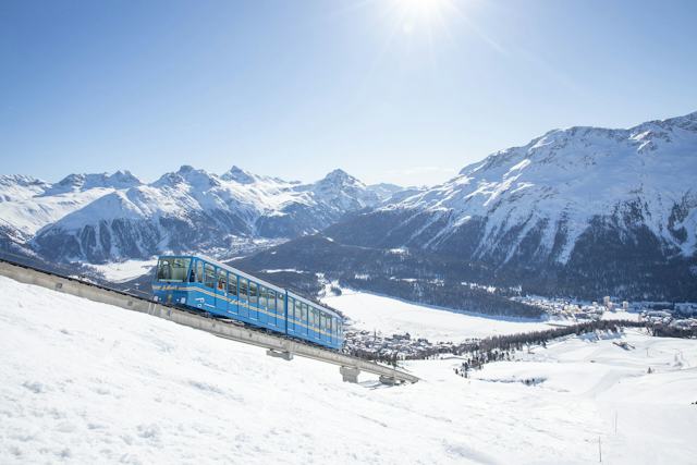 Die blaue Standseilbahn Corviglia mit der Aufschrift «St. Moritz» fährt durch eine verschneite Berglandschaft. Im Hintergrund sind der zugefrorene See, St. Moritz und weitere verschneite Berggipfel zu sehen.