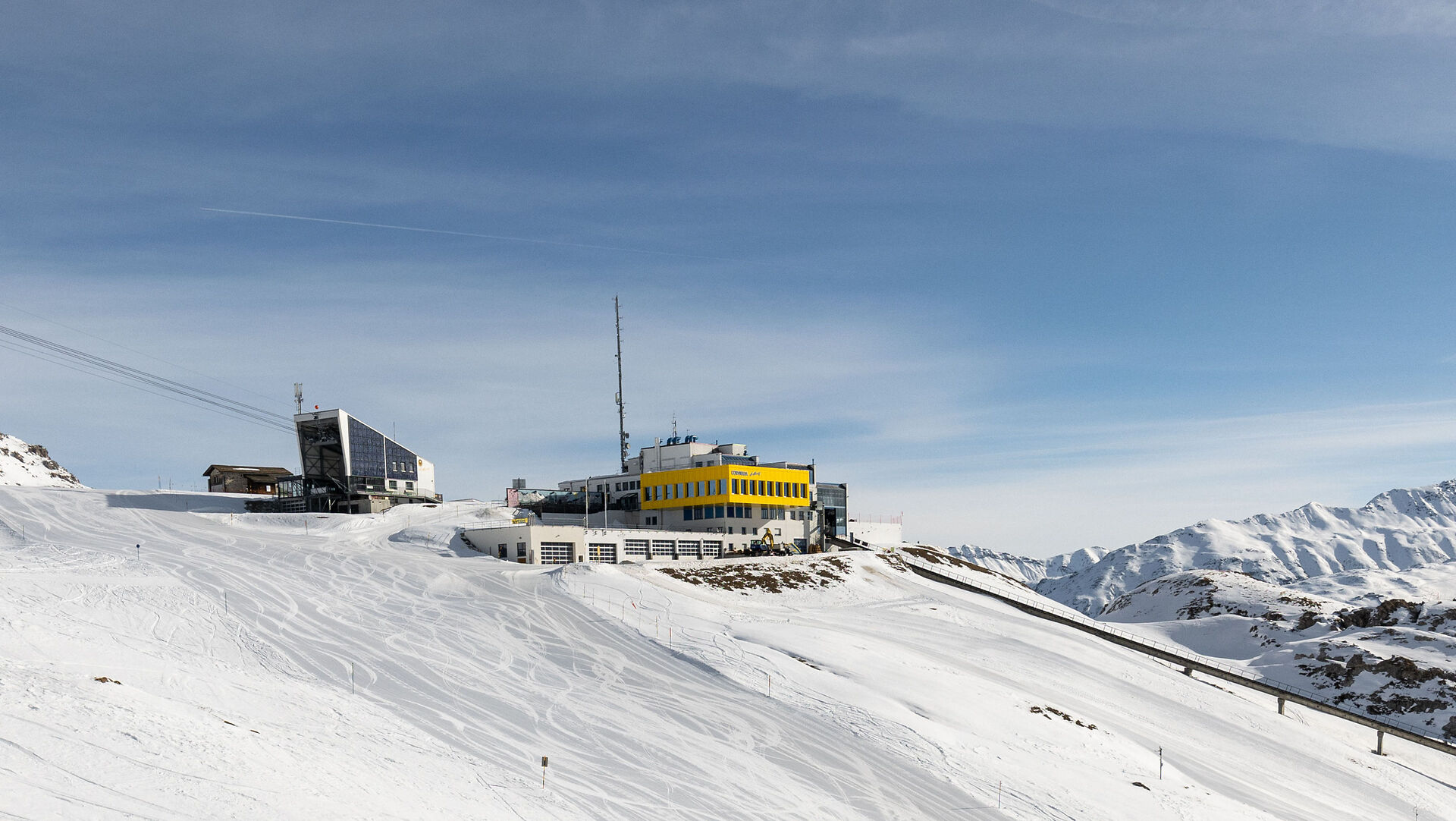 Verschneite Berglandschaft auf Corviglia mit der Seilbahnanlage der Luftseilbahn Piz Nair links im Bild und dem gelben Gebäude der Bergstation Corviglia im Mittelpunkt. Im Hintergrund erstreckt sich eine weitläufige Bergkette mit schneebedeckten Gipfeln unter einem klaren blauen Himmel.