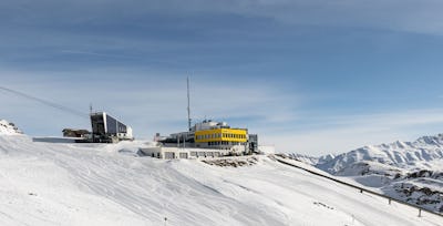 Bergstation Corviglia | Winter Verschneite Berglandschaft auf Corviglia mit der Seilbahnanlage der Luftseilbahn Piz Nair links im Bild und dem gelben Gebäude der Bergstation Corviglia im Mittelpunkt. Im Hintergrund erstreckt sich eine weitläufige Bergkette mit schneebedeckten Gipfeln unter einem klaren blauen Himmel.