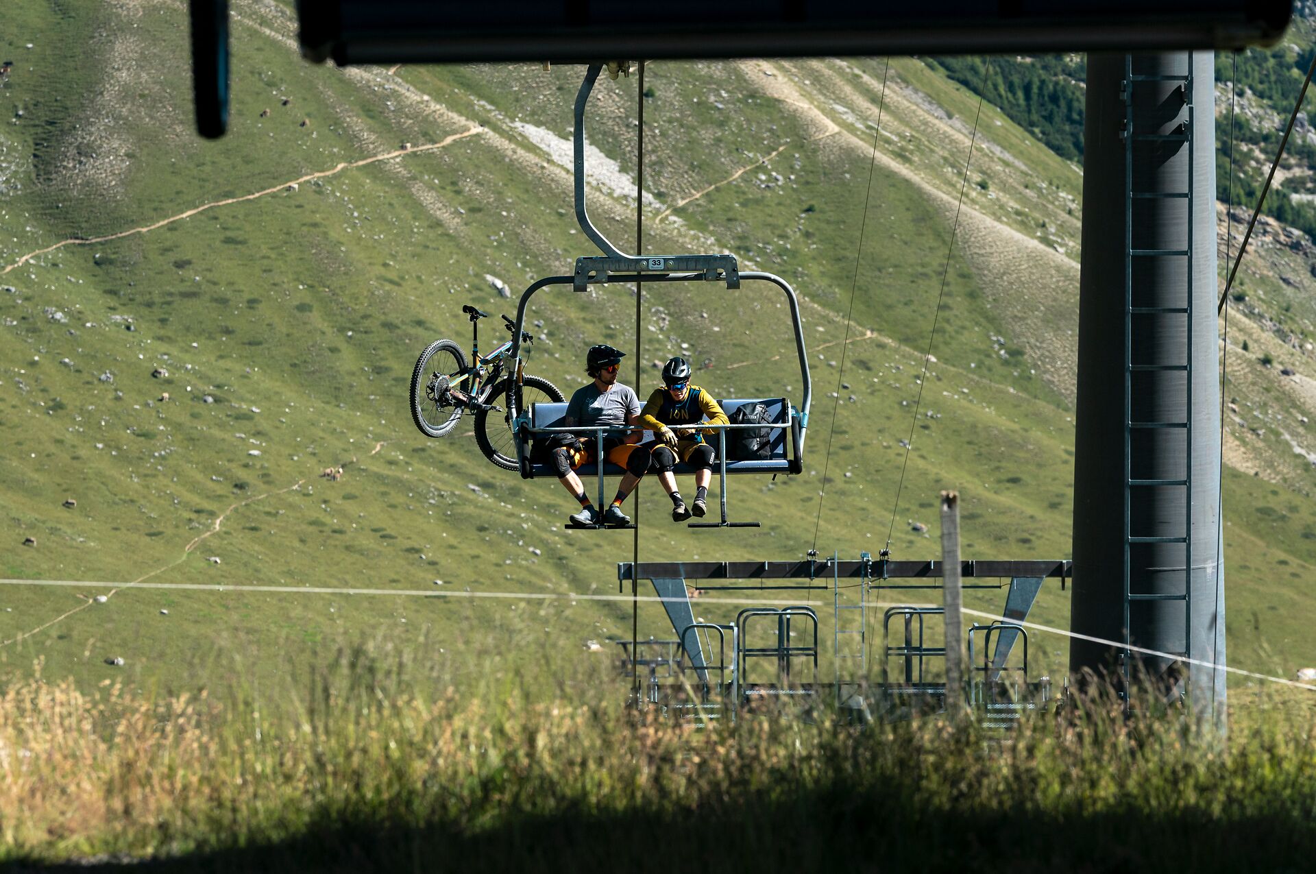 Eine Sesselbahn, an der zwei Mountainbiker mit ihren Rädern befestigt sind, fährt über eine grüne, von Felsen durchzogene Bergwiese.