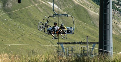 Anlagen | Corviglia | Sommer Eine Sesselbahn, an der zwei Mountainbiker mit ihren Rädern befestigt sind, fährt über eine grüne, von Felsen durchzogene Bergwiese.