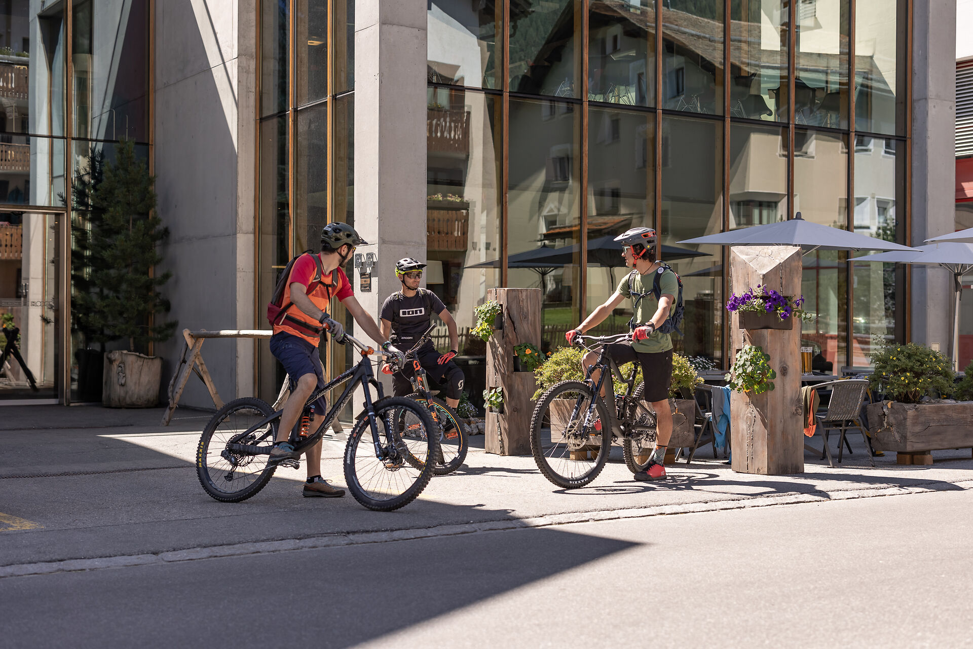 Drei Mountainbiker stehen vordem ALL IN ONE HOTEL INN LODGE auf ihren Fahrrädern und unterhalten sich. Alle drei tragen Helme und Fahrradausrüstung. im Hintergrund befinden sich ein Eingangsbereich mit Glasfront, Holzelemente und Bäume.