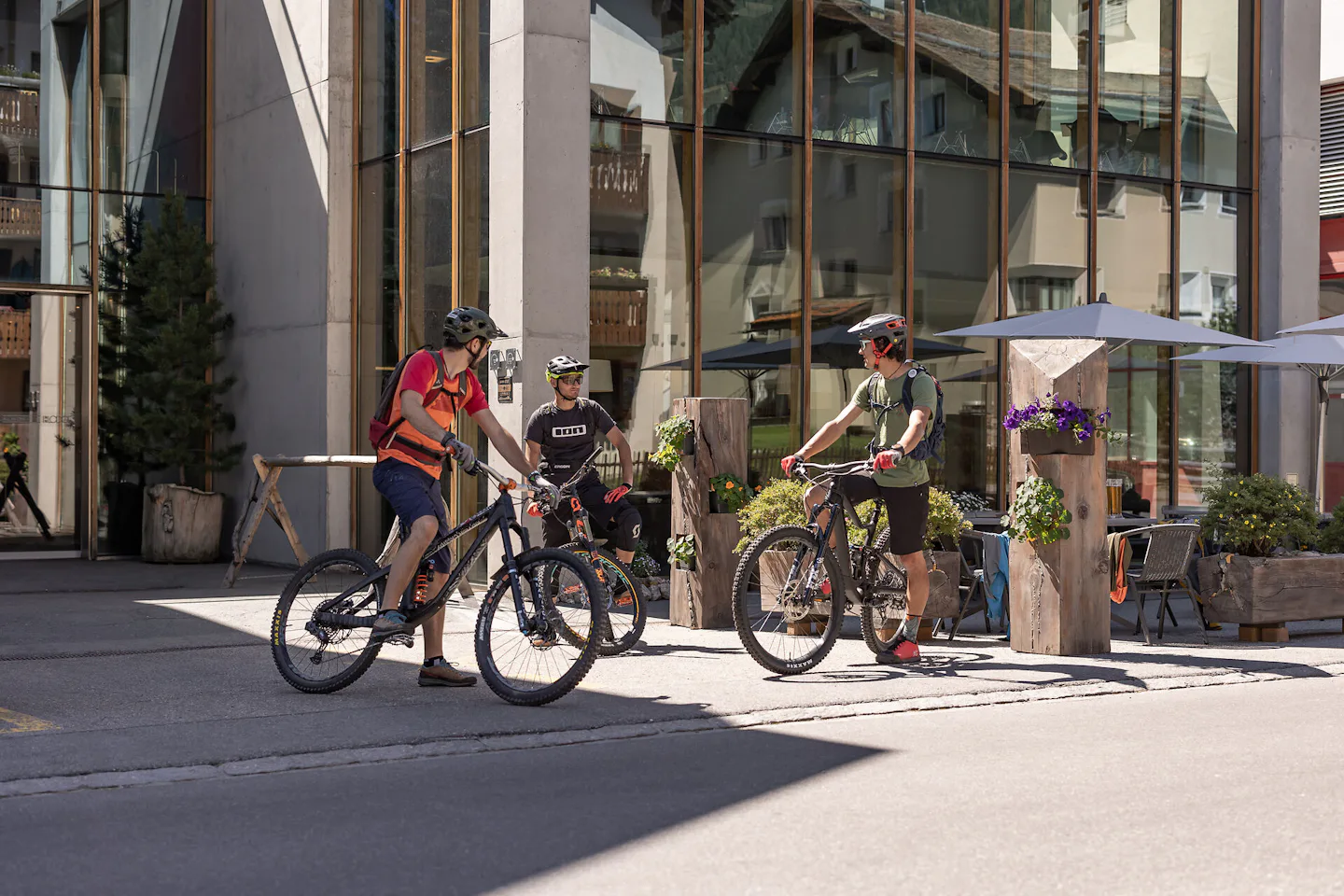 ALL IN ONE HOTEL INN LODGE Celerina | Aussenansicht Sommer Drei Mountainbiker stehen vordem ALL IN ONE HOTEL INN LODGE auf ihren Fahrrädern und unterhalten sich. Alle drei tragen Helme und Fahrradausrüstung. im Hintergrund befinden sich ein Eingangsbereich mit Glasfront, Holzelemente und Bäume.
