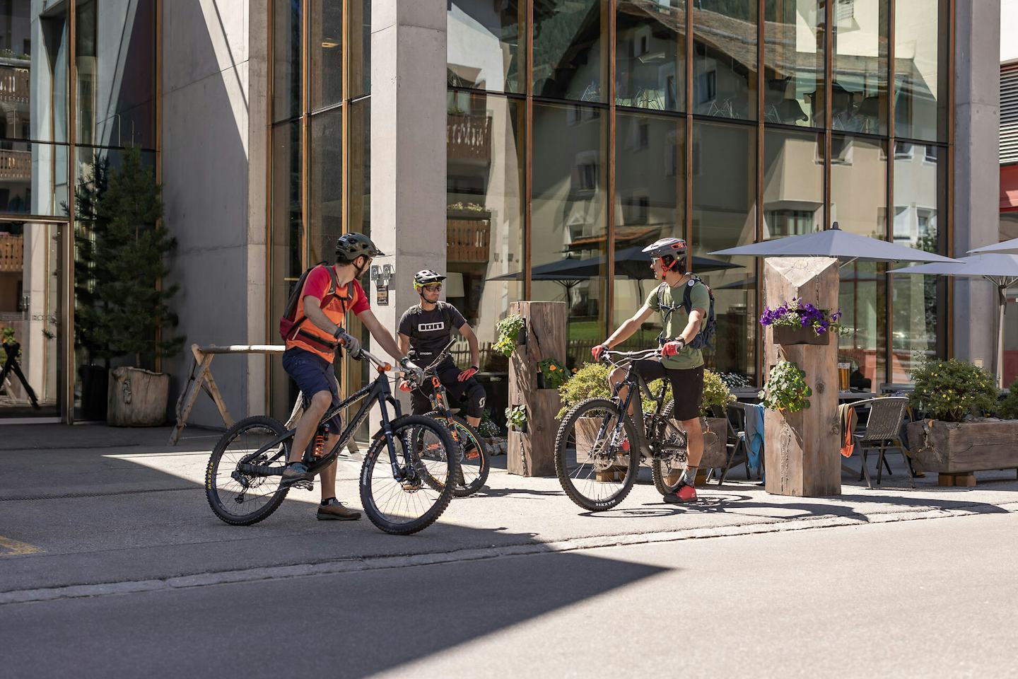 ALL IN ONE HOTEL INN LODGE Celerina | Aussenansicht Sommer Drei Mountainbiker stehen vordem ALL IN ONE HOTEL INN LODGE auf ihren Fahrrädern und unterhalten sich. Alle drei tragen Helme und Fahrradausrüstung. im Hintergrund befinden sich ein Eingangsbereich mit Glasfront, Holzelemente und Bäume.