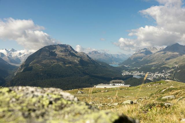 Panoramaansicht des Romantik Hotels Muottas Muragl. Im Hintergrund erstrecken sich bewaldete Berghänge, mehrere teilweise verschneite Berggipfel und ein See; rechts ist ein Teil des Engadiner Tals mit Gebäuden zu sehen.