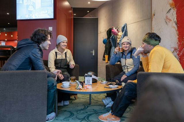 Zwei Skifahrerinnen und zwei Skifahrer sitzen gemeinsam in der Lounge auf grauen Sesseln und trinken warme Getränke.