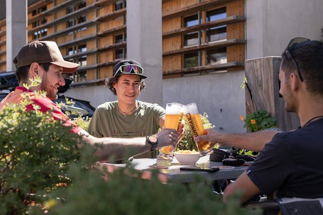Drei Biker sitzen gemeinsam auf der Sonnenterrasse des ALL IN ONE HOTEL INN LODGE  und stossen gemeinsam mit Bier an. Im Hintergrund sind die Holzfenster des Hotels zu sehen und im Vordergrund grüne Bepflanzung.