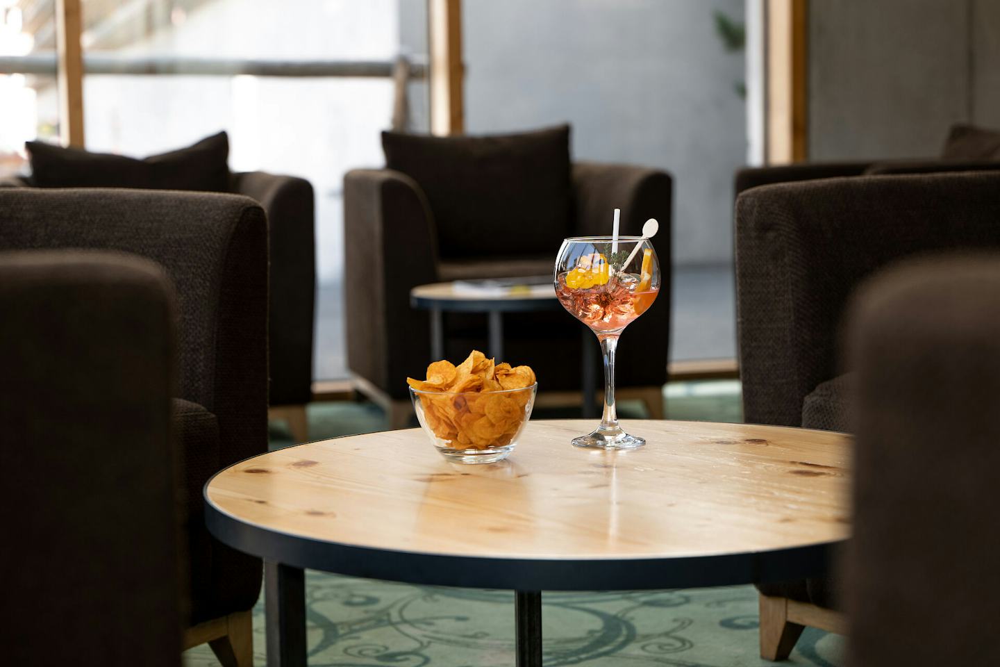 Cocktail mit rotfarbenem Getränk in einem grossen Stielglas auf einem Holztisch und eine Glasschale mit Chips. Im Hintergrund sind Graue Sessel.