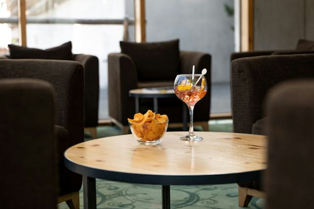 Cocktail mit rotfarbenem Getränk in einem grossen Stielglas auf einem Holztisch und eine Glasschale mit Chips. Im Hintergrund sind Graue Sessel.