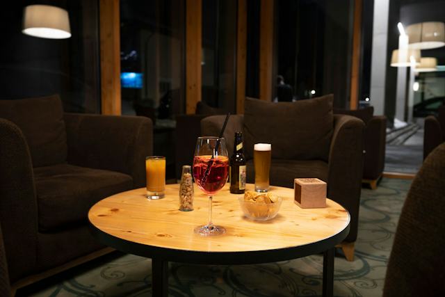 Auf einem Tisch stehen ein roter und ein oranger Cocktail sowie eine Bierflasche mit gefülltem Bierglas und Snacks. Alles ist von roten Sesseln umgeben und es herrscht eine gemütliche Atmosphäre. Draußen ist es dunkel.
