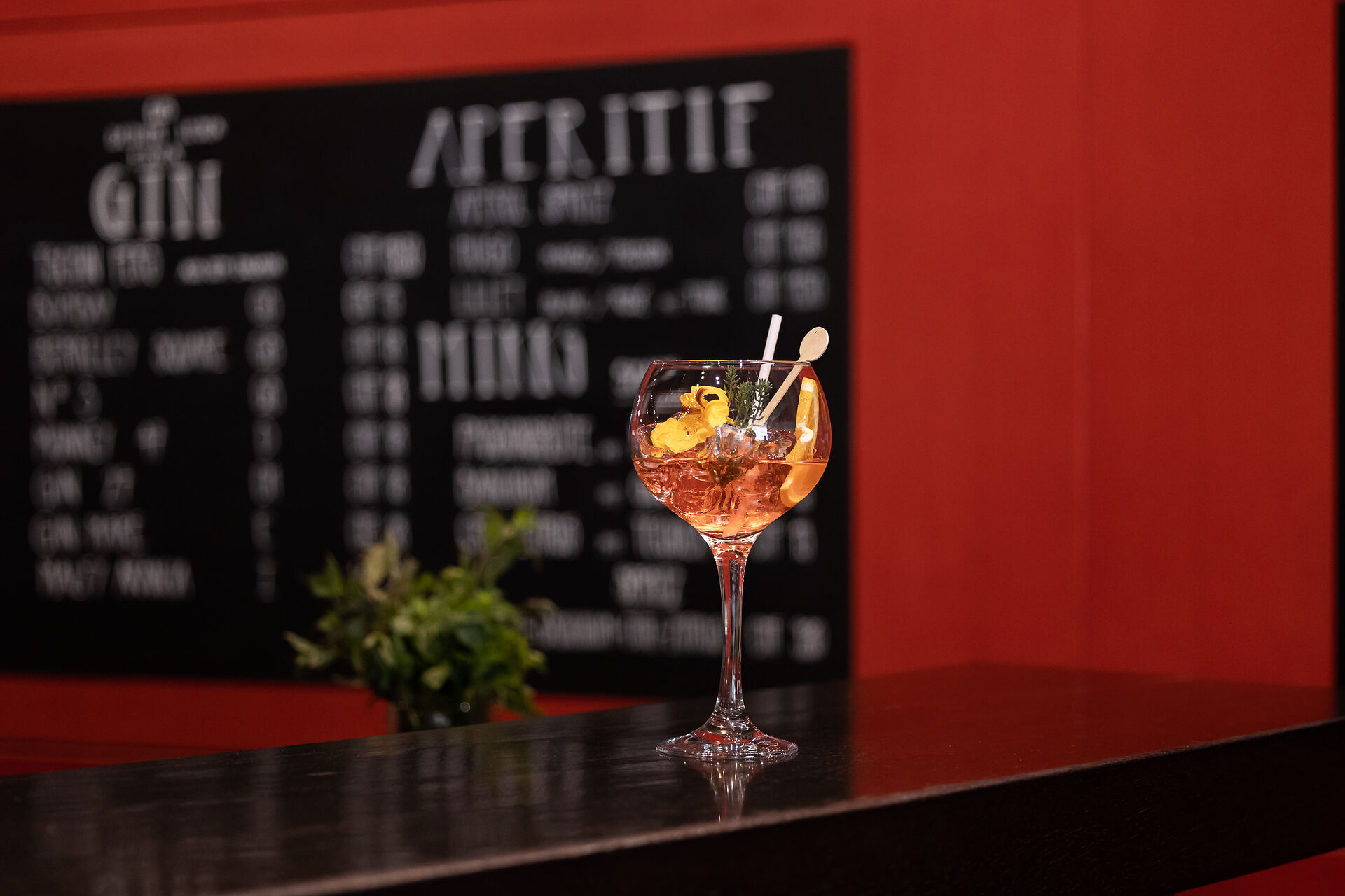 Cocktail mit orangefarbenem Getränk in einem grossen Stielglas auf einer schwarzen Bartheke. Das Getränk ist mit Orangenscheiben, Kräutern und einer dekorativen Blüte garniert. Im Hintergrund sind eine schwarze Getränkekarte und eine rote Wand zu sehen.