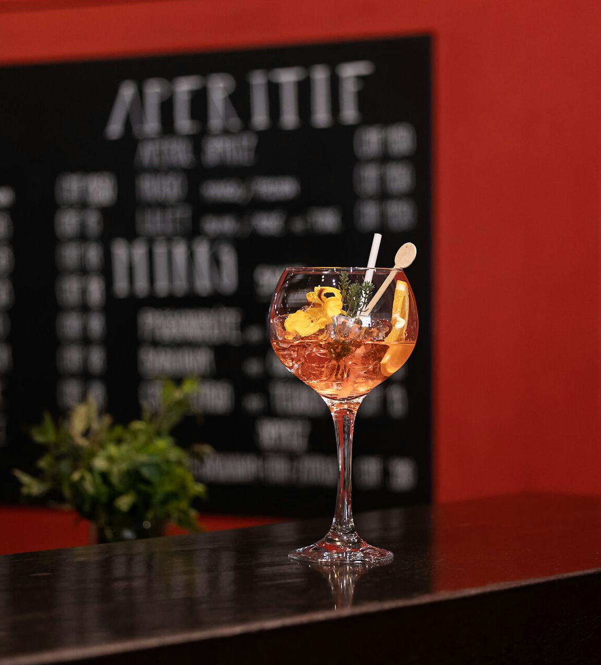 ALL IN ONE HOTEL INN LODGE Celerina | Lounge & Bar Cocktail mit orangefarbenem Getränk in einem grossen Stielglas auf einer schwarzen Bartheke. Das Getränk ist mit Orangenscheiben, Kräutern und einer dekorativen Blüte garniert. Im Hintergrund sind eine schwarze Getränkekarte und eine rote Wand zu sehen.