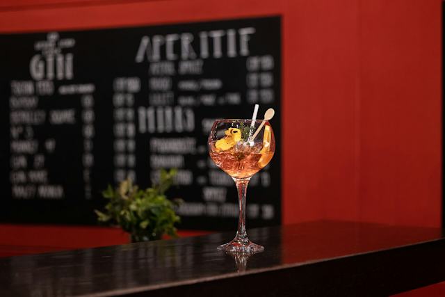 Cocktail mit orangefarbenem Getränk in einem grossen Stielglas auf einer schwarzen Bartheke. Das Getränk ist mit Orangenscheiben, Kräutern und einer dekorativen Blüte garniert. Im Hintergrund sind eine schwarze Getränkekarte und eine rote Wand zu sehen.