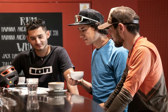 Drei Mountainbiker geniessen einen Kaffee während sie auf eine Karte auf einer schwarzen Bartheke schauen. Das Getränk ist mit Orangenscheiben, Kräutern und einer dekorativen Blüte garniert. Im Hintergrund sind eine schwarze Getränkekarte und eine rote Wand zu sehen.