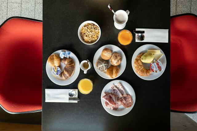 Frühstückstisch im ALL IN ONE HOTEL INN LODGE mit drei Tellern aus der Vogelperspektive. Auf dem Tisch stehen Gläser mit Orangensaft, Tassen, eine Schale mit Müsli, eine Birne, Äpfel sowie Teller mit Brot, Käse und Schinken.