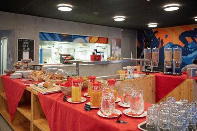 Frühstücksbuffet im ALL IN ONE HOTEL INN LODGE mit verschiedenen Backwaren, Brot, Äpfeln und weiteren Früchten, Müsli- beziehungsweise Cerealienbehältern sowie mehreren Gläsern mit Getränken. Auf der Theke stehen zudem Tassen, Teller, Besteck und weitere Frühstücksutensilien. Im Hintergrund ist die Küche und eine grossflächige farbige Wandgestaltung mit Bergmotiven und Wintersportlern zu sehen.