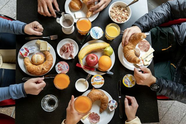 Frühstückstisch im ALL IN ONE HOTEL INN LODGE mit vier Personen aus der Vogelperspektive. Auf dem Tisch stehen Gläser mit Orangensaft, Tassen, eine Schale mit Müsli, eine Birne, Äpfel sowie Teller mit Brot, Käse und Schinken.