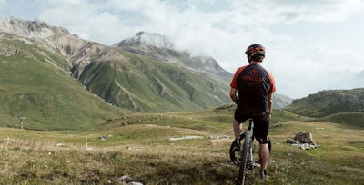 Mountainbike | Corviglia | Sommer Ein in schwarz-roter Montur gekleideter Mountainbiker sitzt auf seinem Bike und schaut auf die grüne Berglandschaft von Corviglia.