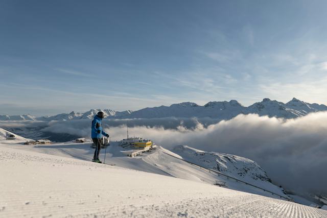 Auf einer frisch präparierten Skipiste steht ein Skifahrer mit blauer Jacke. Im Hintergrund sind die gelbe Bergstation Corviglia und das im Nebel liegende Tal zu sehen, während darüber die verschneiten Gipfel der Engadiner Bergwelt hinausragen.