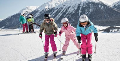 Familienangebote | Corviglia | Winter Drei Kinder fahren Ski und eine Mutter und zwei Kinder stehen auf Skiern im Hintergrund und schauen ins Tal. Sie tragen farbige Winterbekleidung und Skibrillen beziehungsweise Helme. Im Hintergrund ist eine weitläufige verschneite Berglandschaft mit bewaldeten Hängen und mehreren Berggipfeln zu sehen.