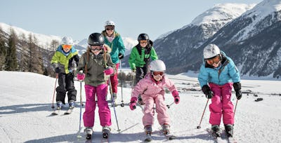 Familienangebote | Corviglia | Winter Fünf Kinder und Jugendliche und eine Mutter fahren Ski. Sie tragen farbige Winterbekleidung und Skibrillen beziehungsweise Helme. Im Hintergrund ist eine weitläufige verschneite Berglandschaft mit bewaldeten Hängen und mehreren Berggipfeln zu sehen.