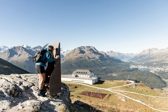 Auf dem Bild ist eine Wanderin zu sehen, die ins Engadiner Tal mit seinen Seen schaut. In der Bildmitte ist das Romantik Hotel Muottas Muragl zu erkennen.