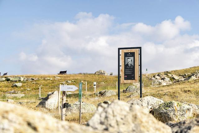 Blick über die alpine Landschaft auf Muottas Muragl mit einer Informationstafel des Philosophenwegs im Vordergrund. Die Tafel steht auf einer grasbewachsenen, felsigen Fläche neben einem Weg und zeigt ein Porträt sowie Text.