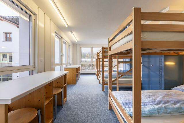 Helle 6-Bettzimmer des ALL IN ONE HOTEL INN LODGE mit mehreren hölzernen Etagenbetten, die entlang der rechten Seite des Raumes angeordnet sind. Die Betten sind mit weisser Bettwäsche bezogen. Entlang der linken Wand befinden sich mehrere Fenster sowie lange, helle Ablageflächen mit integrierten Sitzplätzen. Im hinteren Bereich stehen weitere hölzerne Schränke und Ablagen. Durch die Fenster fällt Tageslicht in den Raum; durch die grossen Fenster im hinteren Bereich ist eine winterliche Landschaft zu sehen.