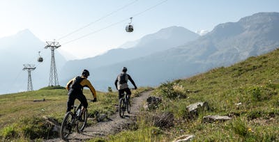 Mountainbike | Corviglia | Sommer Zwei Mountainbiker fahren auf einem schmalen, steinigen Bergweg durch eine alpine Landschaft. Beide tragen Helme und Rucksäcke; der vordere Fahrer trägt eine schwarze und weisse Bekleidung, der hintere eine schwarze Hose und ein gelbes Oberteil. Oberhalb des Weges verlaufen mehrere Seilbahnkabel mit daran hängenden Gondeln. Im Hintergrund erstreckt sich eine weitläufige Berglandschaft mit bewaldeten Hängen und hohen Berggipfeln unter einem hellen Himmel.