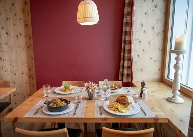 Panoramarestaurant | Romantik Hotel Muottas Muragl Holztisch im Panoramarestaurant Muottas Muragl mit vierangerichteten Gerichten. Im Vordergrund steht eine schwarze Schale mit einem Gericht und knusprigen Zwiebeln, daneben Besteck und ein Glas. Auf den weiteren Tellern sind ein Gericht mit einer hellen Beilage sowie ein Burger mit Pommes frites zu sehen. Auf dem Tisch stehen mehrere Wassergläser, eine Wasserkaraffe, Gewürzmühlen und eine Vase mit getrockneten Blumen. Im Hintergrund sind ein Fenster und rot-weiss karierte Vorhänge zu sehen.