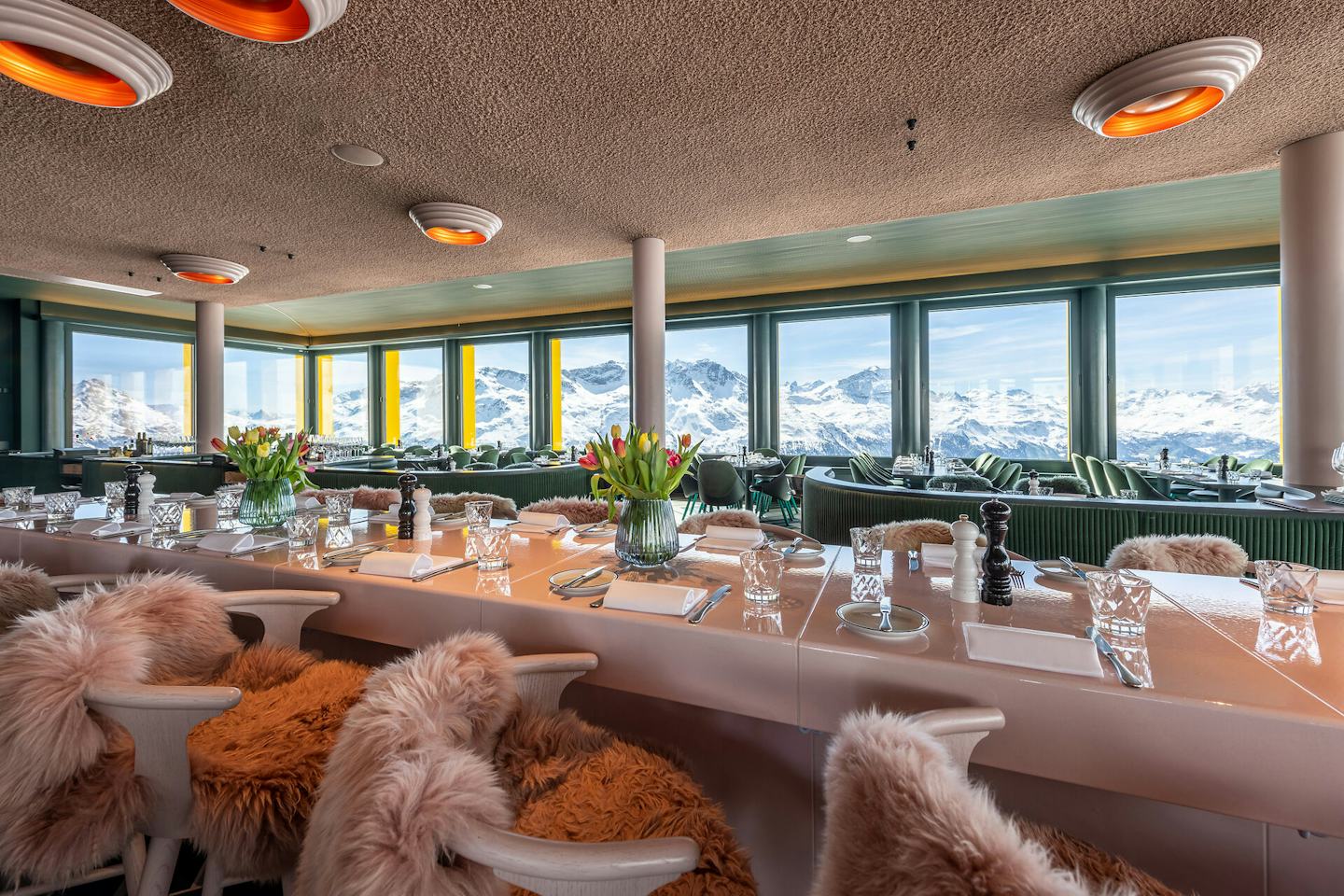 Restaurant White Marmot | Corviglia | Winter Heller Innenraum des White Marmot im Skigebiet Corviglia mit mehreren langen, gedeckten Tischen und Sitzplätzen. Die Tische sind mit Besteck, Gläsern, Servietten, Gewürzmühlen und Blumenvasen dekoriert. Die weissen Stühle sind teilweise mit flauschigen Fellauflagen versehen. Entlang der Fensterfront befinden sich weitere Sitzplätze mit dunkelgrünen Polsterungen. Durch die grossen Fenster ist eine weitläufige verschneite Berglandschaft mit zahlreichen Berggipfeln zu sehen. An der Decke hängen mehrere runde Leuchten mit orangefarbenen Innenflächen.