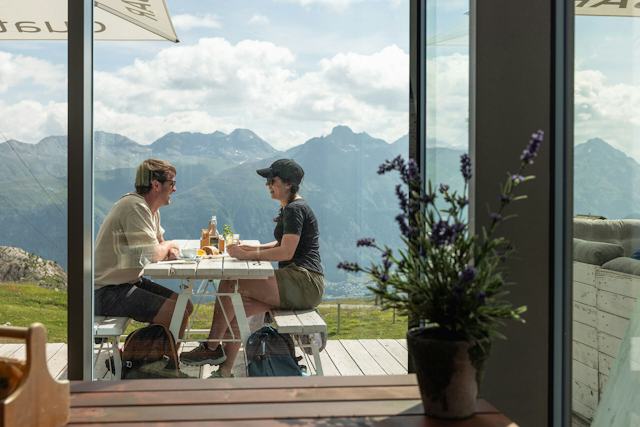 quattro BAR | Corviglia | Sommer Sonnenterrasse der Quattro Bar mit zwei Personen. Im Vordergrund ein Holztisch und im Hintergrund befinden sich grosse Fenster des Gebäudes und eine weitläufige Berglandschaft unter blauem Himmel mit ein Wolken.