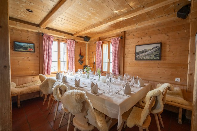 Rustikaler Gastraum der Villa Lyss mit holzverkleideten Wänden und Decke. Ein grosser Tisch ist mit weisser Tischdecke, gefalteten Servietten, Besteck sowie zahlreichen Wein- und Wassergläsern festlich gedeckt. Die Holzstühle und Sitzbänke sind mit weissen Fellen bedeckt. An den Fenstern hängen rot-weiss karierte Vorhänge; an den Wänden sind gerahmte Bilder zu sehen.