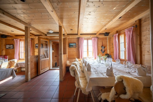 Rustikal eingerichteter Gastraum der Villa Lyss mit holzverkleideten Wänden und Decke, sichtbaren Holzbalken und rot-weiss karierten Vorhängen. Mehrere Tische sind mit weissen Tischdecken, gefalteten Servietten, Besteck sowie zahlreichen Wein- und Wassergläsern gedeckt. Die Stühle sind mit weissen Fellen bedeckt. An den Wänden hängen gerahmte Bilder und dekorative Elemente; durch die Fenster fällt Tageslicht in den Raum.