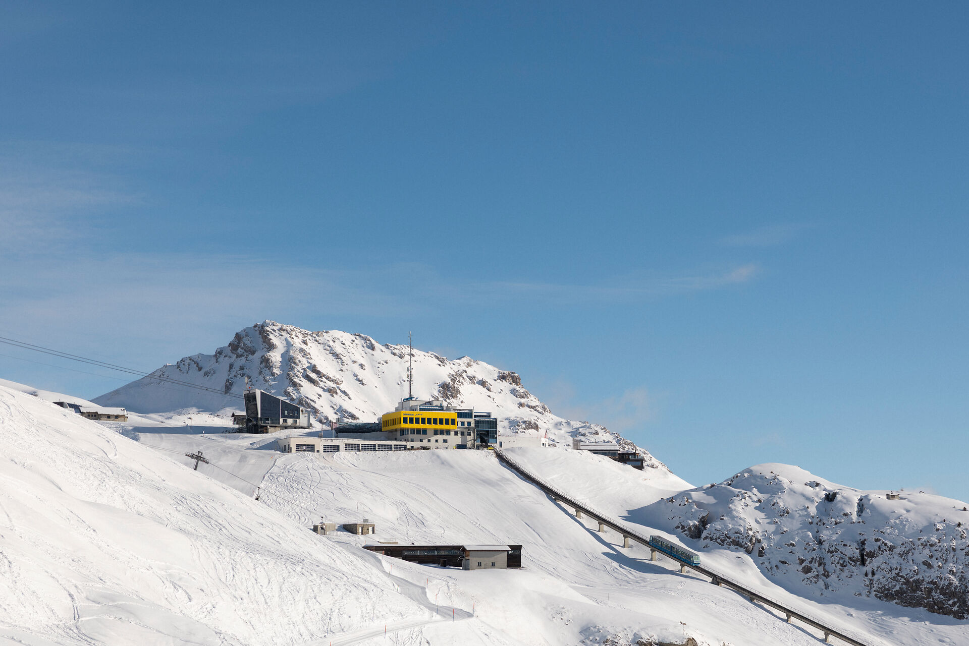 Die Aufnahme zeigt die gelbe Bergstation Corviglia mit ihren weissen Pisten im Vordergrund und einem blauen Himmel.