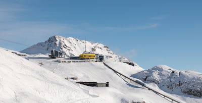 Bergstation Corviglia | Winter Die Aufnahme zeigt die gelbe Bergstation Corviglia mit ihren weissen Pisten im Vordergrund und einem blauen Himmel.