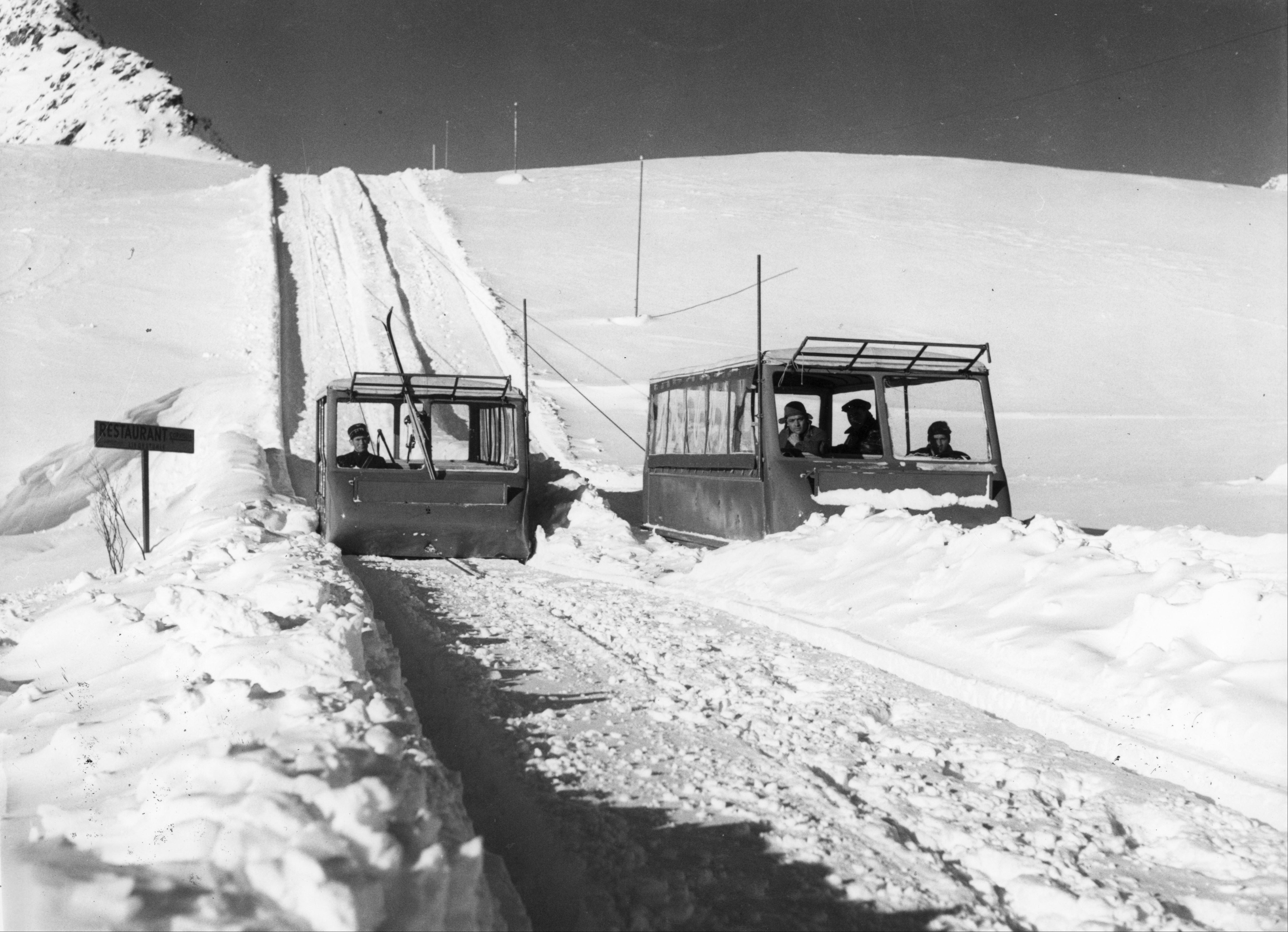 Historische Schwarz-Weiss-Aufnahme von der ersten Standseilbahn auf Corviglia. In den Fahrzeugen sitzen mehrere Personen. Links neben der Strasse steht ein Wegweiser mit der Aufschrift «RESTAURANT». Im Hintergrund führt die Strecke einen steilen, verschneiten Hang hinauf; entlang der Strecke sind mehrere Masten und Leitungen zu sehen.