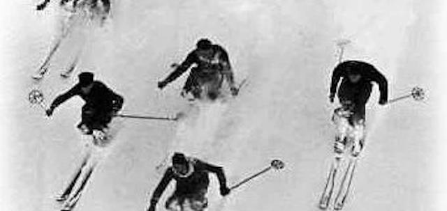 Historische Schwarz-Weiss-Aufnahme von vier Skifahrern bei der Abfahrt auf einem verschneiten Hang. Die Personen fahren mit Skiern und Skistöcken in unterschiedlicher Position den Hang hinunter; zwei befinden sich im Vordergrund, zwei weitere dahinter.