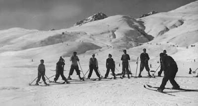 Historische Schwarz-Weiss-Aufnahme von mehreren Skifahrern auf einem verschneiten Berghang der Corviglia. Die Personen stehen mit Skiern und Skistöcken im Schnee, während im Vordergrund ein Skifahrer den Hang hinunterfährt. Im Hintergrund sind sanft ansteigende, schneebedeckte Bergflanken und mehrere Berggipfel zu sehen.