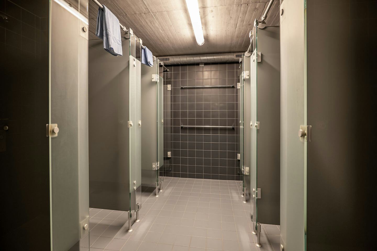 Gemeinschaftlicher Duschbereich im ALL IN ONE HOTEL INN LODGE mit mehreren abgetrennten Duschen und geöffneten Türen. Der Raum ist mit dunkelgrauen Wandfliesen und grauen Bodenfliesen ausgestattet; an der hinteren Wand befinden sich mehrere horizontale Handläufe. An der Decke verlaufen sichtbare Leitungen und lange Leuchten.