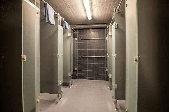 Gemeinschaftlicher Duschbereich im ALL IN ONE HOTEL INN LODGE mit mehreren abgetrennten Duschen und geöffneten Türen. Der Raum ist mit dunkelgrauen Wandfliesen und grauen Bodenfliesen ausgestattet; an der hinteren Wand befinden sich mehrere horizontale Handläufe. An der Decke verlaufen sichtbare Leitungen und lange Leuchten.