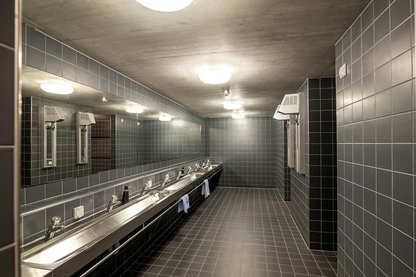 Sanitärbereich | ALL IN ONE HOTEL INN LODGE Moderner Waschbereich im ALL IN ONE HOTEL INN LODGE mit grauen Wand- und Bodenfliesen, einer langen Waschtischanlage mit mehreren Waschbecken und Armaturen sowie einem grossen Spiegel über der gesamten Waschanlage. An der Wand befinden sich ein Händetrockner und Handtuchhalter; mehrere runde Deckenleuchten erhellen den Raum.
