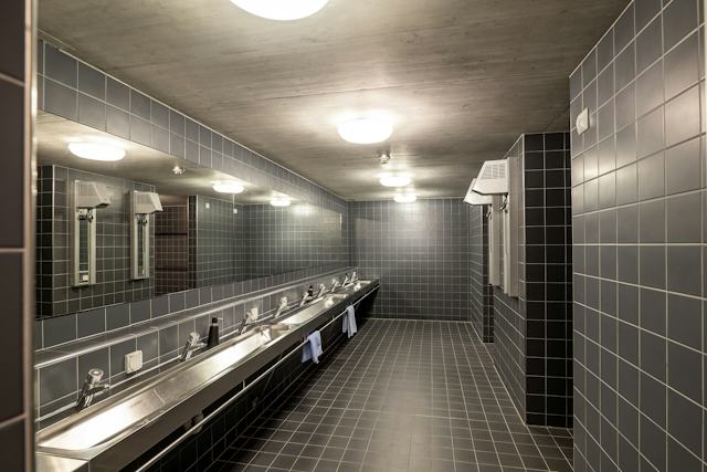 Moderner Waschbereich im ALL IN ONE HOTEL INN LODGE mit grauen Wand- und Bodenfliesen, einer langen Waschtischanlage mit mehreren Waschbecken und Armaturen sowie einem grossen Spiegel über der gesamten Waschanlage. An der Wand befinden sich ein Händetrockner und Handtuchhalter; mehrere runde Deckenleuchten erhellen den Raum.