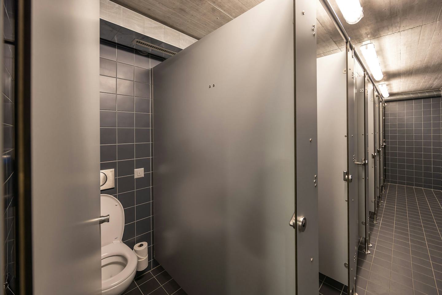 Sanitärbereich | ALL IN ONE HOTEL INN LODGE Moderner Sanitärbereich des ALL IN ONE HOTEL INN LODGE mit grauen Türen, dunklen Wand- und Bodenfliesen sowie mehreren abgetrennten Toilettenkabinen. Im Vordergrund ist eine geöffnete Toilettenkabine mit WC und Toilettenpapier zu sehen.