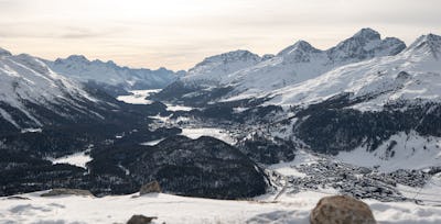 Landscape | Muottas Muragl | Winter Diese Panoramaaufnahme zeigt Muottas Muragl und die verschneite Engadiner Berglandschaft im Winter. Zu erkennen sind bewaldete Berghänge, mehrere verschneite Berggipfel sowie ein Tal mit Siedlungen und Verkehrswegen. Im Vordergrund befindet sich ein breiter, verschneiter Hang mit Steinen. Über der Berglandschaft erstreckt sich ein teilweise bewölkter, blauer Himmel kurz vor Sonnenuntergang.