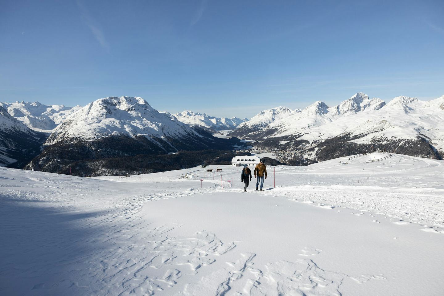 Winterwandern| Muottasmuragl Bergerlebnis | Winter Auf einem verschneiten Winterwanderweg auf Muottas Muragl wandert ein Paar. Eine Person trägt eine schwarze Jacke, die andere eine braune Winterjacke. Im Hintergrund sind das Romantik Hotel Muottas Muragl und die weitläufige, verschneite Berglandschaft des Engadins mit bewaldeten Hängen, Bergseen und zahlreichen schneebedeckten Gipfeln zu sehen. Im Tal sind Häuser und Siedlungen zu erkennen.
